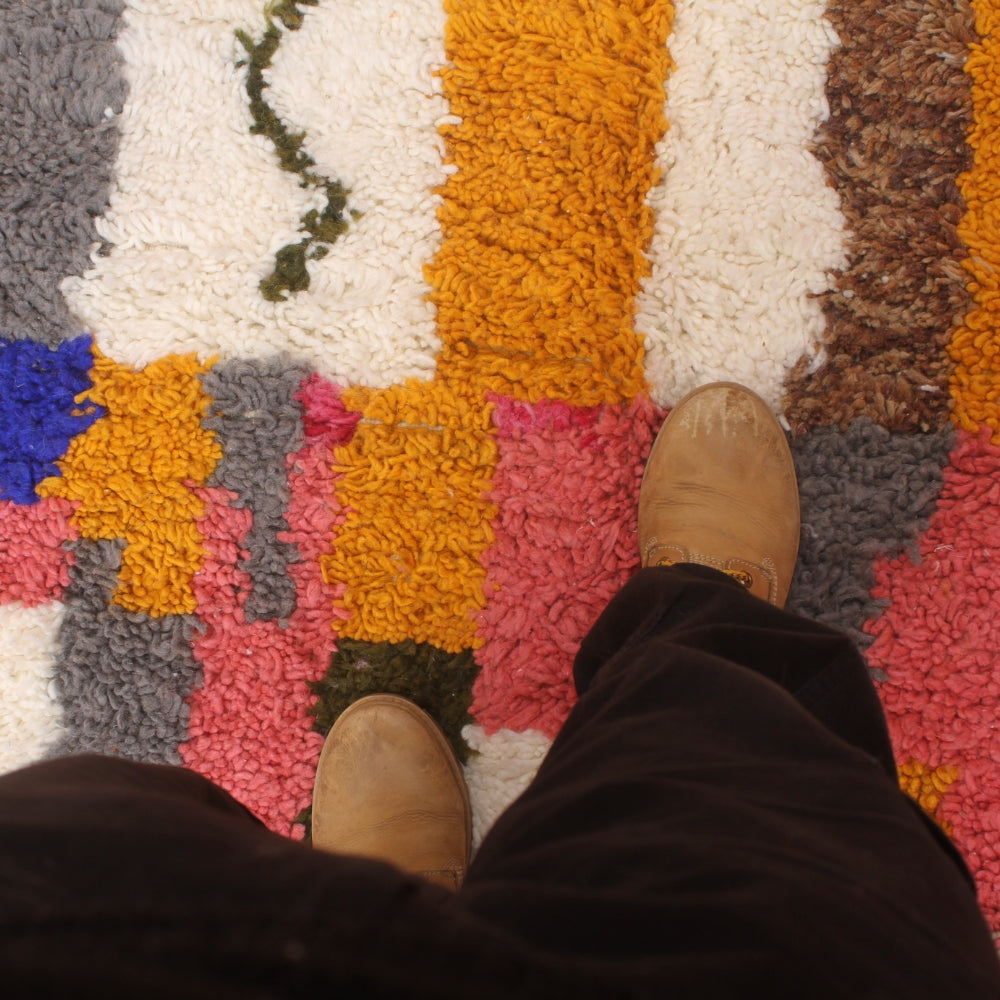 Colorful textured rug with a person's feet wearing brown shoes on a multicolored striped berber rug.